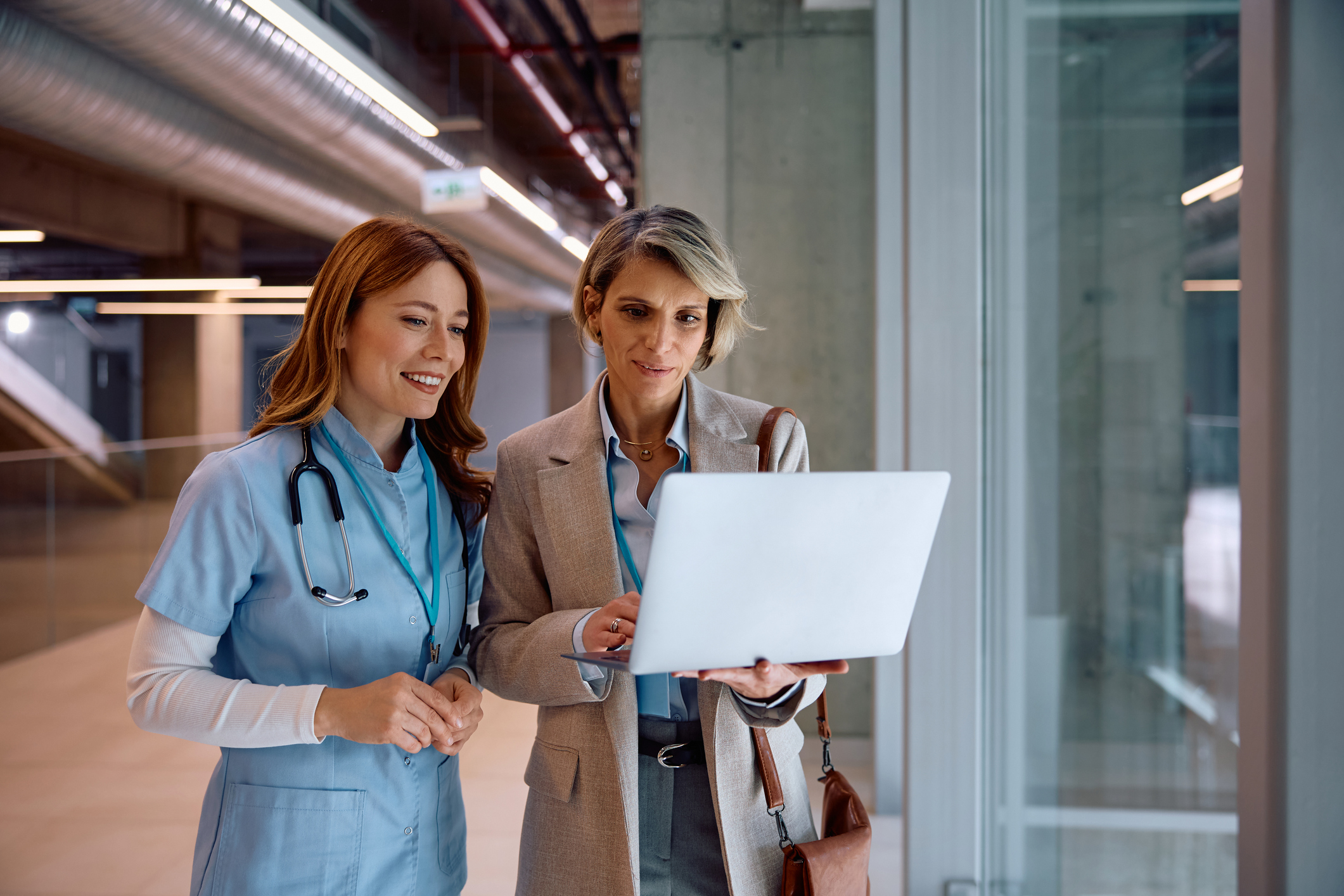 A patient care manager shares a laptop screen with a nurse in a hospital corridor.