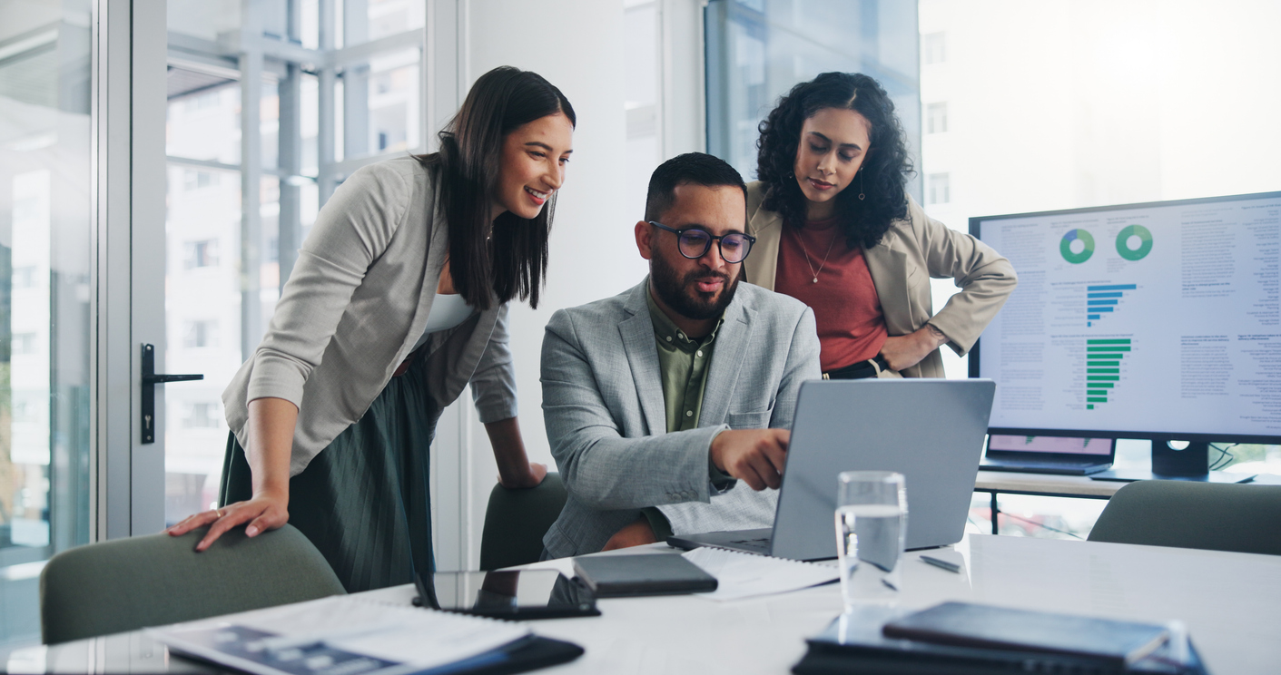Three It Workers Look at a Laptop Screen With a Dashboard on a Monitor Behind Them..jpg