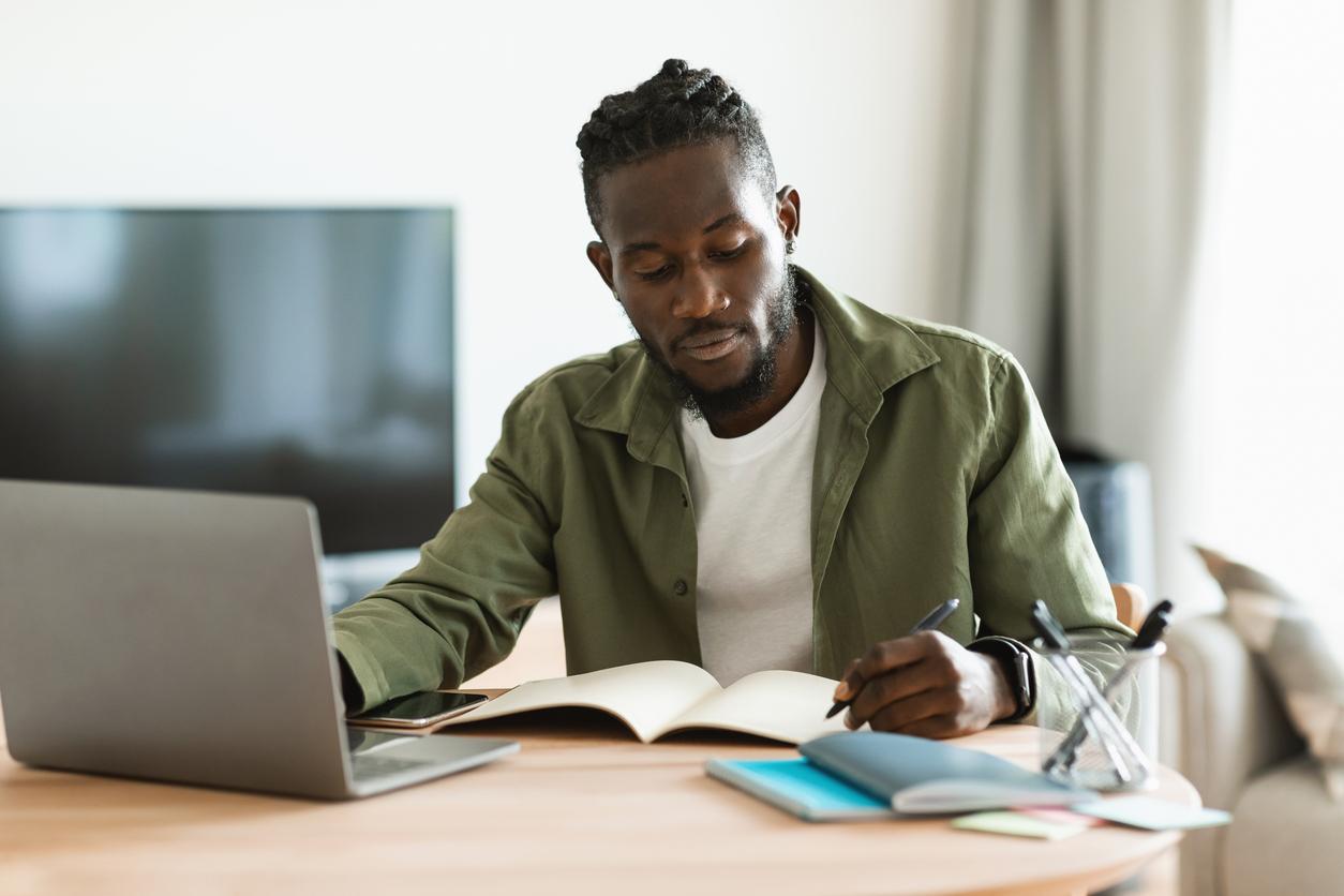 An online business student writes in a notebook while working on a laptop.